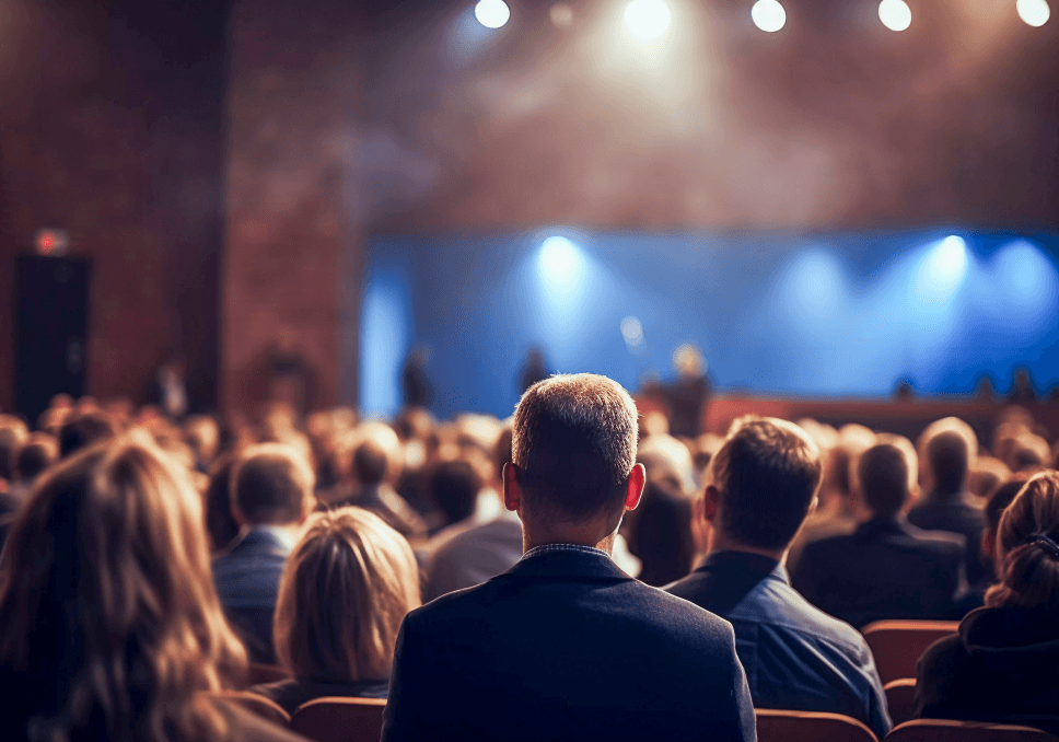 Sur l'image, nous pouvons voir une conférence avec plusieurs personnes assises en train d'écouter les différents interlocuteurs parlés devant eux.