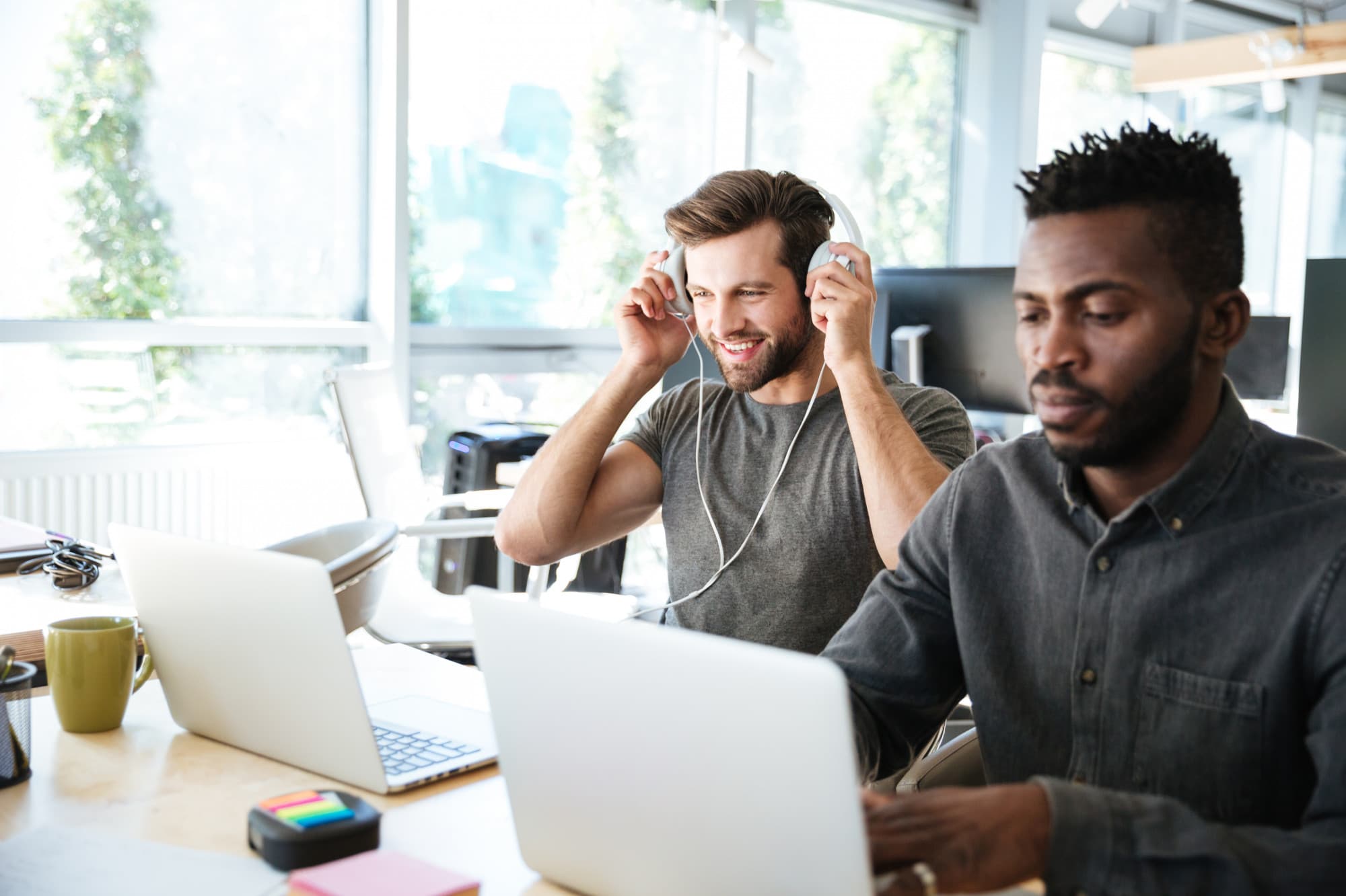 Sur l'image, nous pouvons voir deux personnes derrière un bureau : la personne de gauche positionne son casque et regarde un écran d'ordinateur. La deuxième personne à droite regarde un écran d'ordinateur et tape sur son clavier.